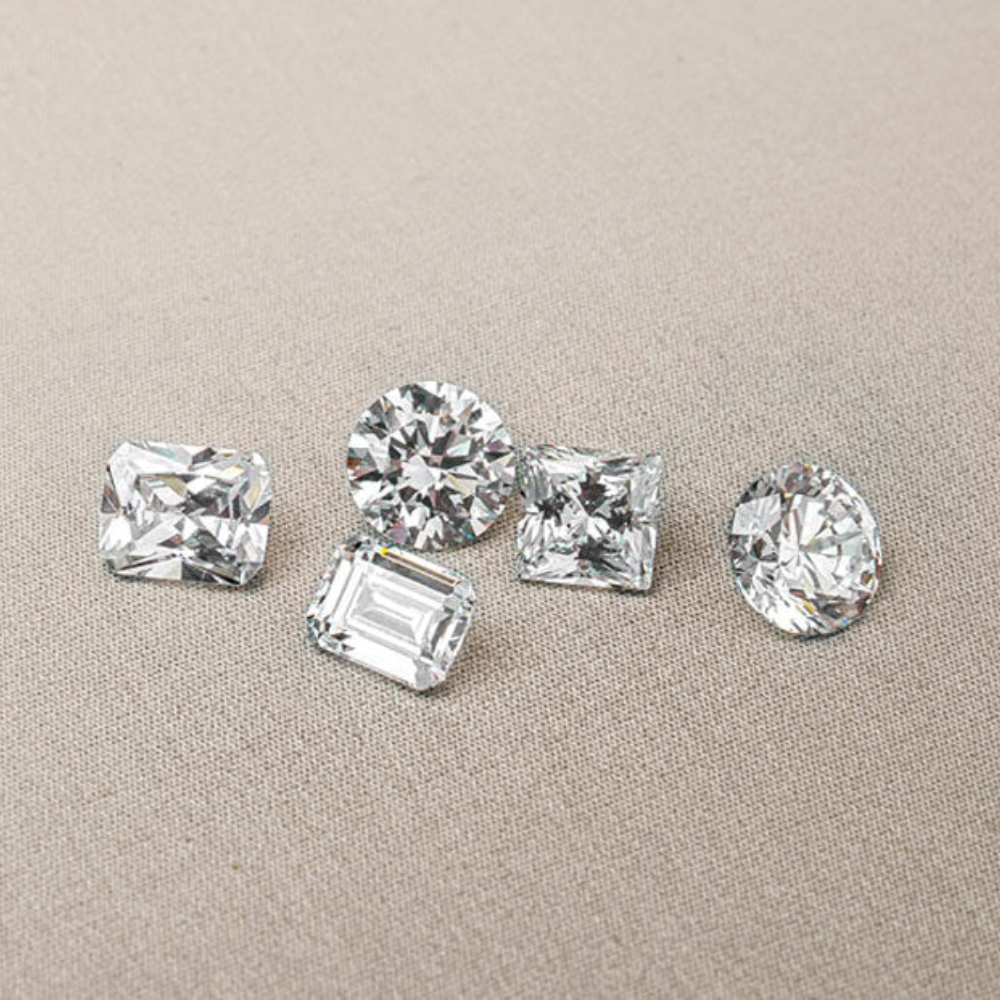 Elegante Präsentation von fünf Diamanten in verschiedenen Schliffen: Smaragd-, Rund-, Kissen-, Prinzess- und Ovalschliff, auf einer neutralen Stoffunterlage. Perfekt geeignet zur Hervorhebung der Vielfalt an Diamantdesigns für Schmuckliebhaber. Optimiert für SEO in den Bereichen Schmuck, Diamanten und Luxusaccessoires.